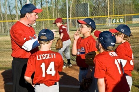 A baseball coach in a red shirt and cap talks to a group of young players, also in red uniforms, at a field.