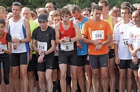 A crowd of runners wearing numbered bibs and athletic gear stand at the starting line of a race, focused and waiting to begin. 