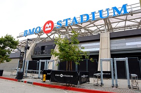 Front view of BMO Stadium entrance with bold blue signage, metal detectors, and a small tree planted nearby.