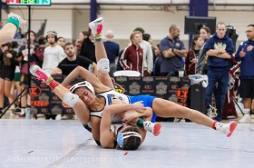 Two female wrestlers in singlets are engaged in a wrestling match on a mat, with one taking down the other as spectators watch in the background.