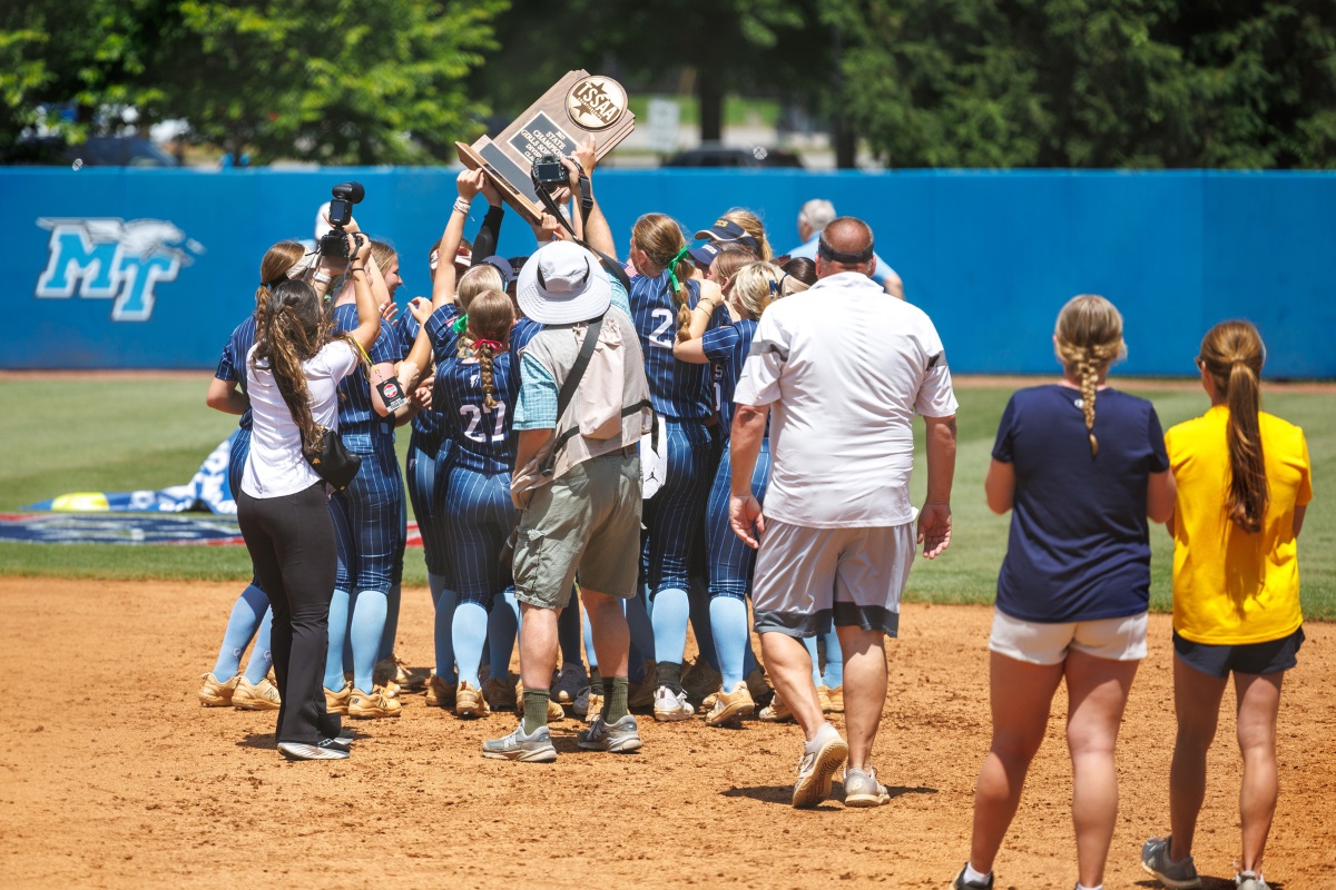 A softball team in blue uniforms celebrates on a sunny field, holding a trophy high. 