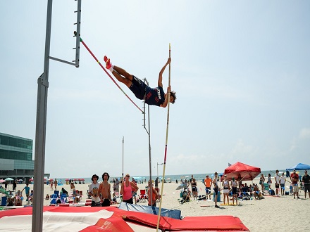 A pole vaulter in mid-air above a beach setting, surrounded by spectators and a coastal backdrop, conveys excitement and athleticism.