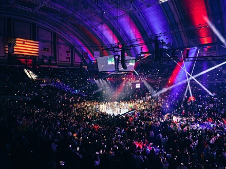 A large crowd fills an indoor arena with a boxing ring at the center, colorful lights overhead, and an American flag on the left wall.