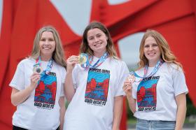 Three smiling women in matching white t-shirts with "Summer Games Grand Rapids MI" printed on them hold up gold, silver, and bronze medals.