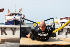 Strongman competitor "Bosman" strains while pulling a thick yellow rope in front of a crowded boat.