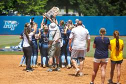 A softball team celebrates with a trophy on a baseball field under a bright sun.