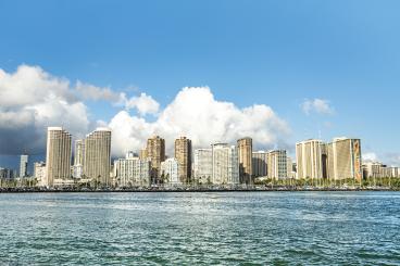 High-rise buildings form a city skyline on a coastline with a blue sky and clouds above a body of water.