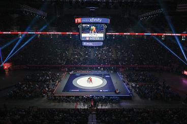 Wrestlers compete on a mat in a large arena during the U.S. Olympic Team Trials, with a crowd watching and a large jumbotron displaying the event.