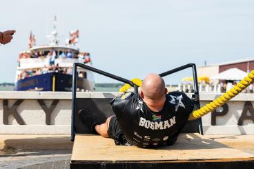 Strongman competitor "Bosman" strains while pulling a thick yellow rope in front of a crowded boat.