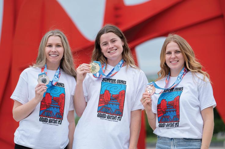 Three smiling women in matching white t-shirts with "Summer Games Grand Rapids MI" printed on them hold up gold, silver, and bronze medals.