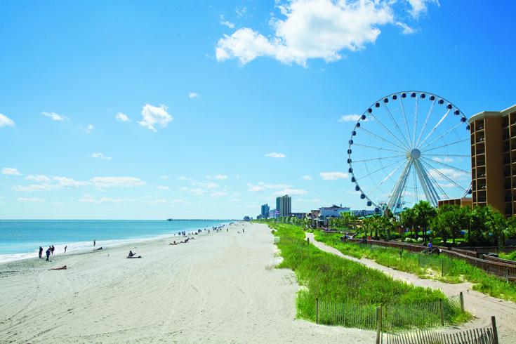 A wide sandy beach lines the ocean under a bright blue sky with scattered clouds, featuring a large Ferris wheel and beachside buildings.