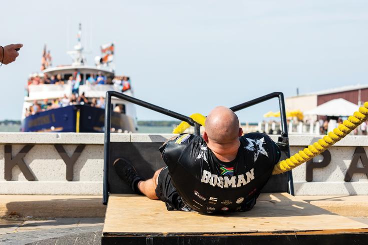 Strongman competitor "Bosman" strains while pulling a thick yellow rope in front of a crowded boat.