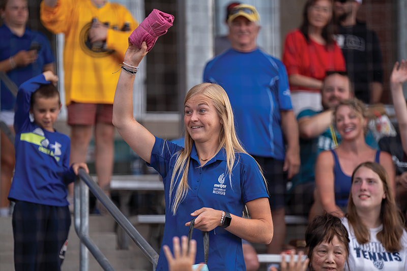 A young woman in a blue polo shirt with a Meijer logo waves a pink folded cloth at an outdoor event, surrounded by spectators.
