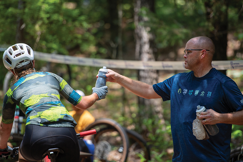 A man in a blue t-shirt hands a water bottle to a cyclist wearing a white helmet and a patterned jersey.