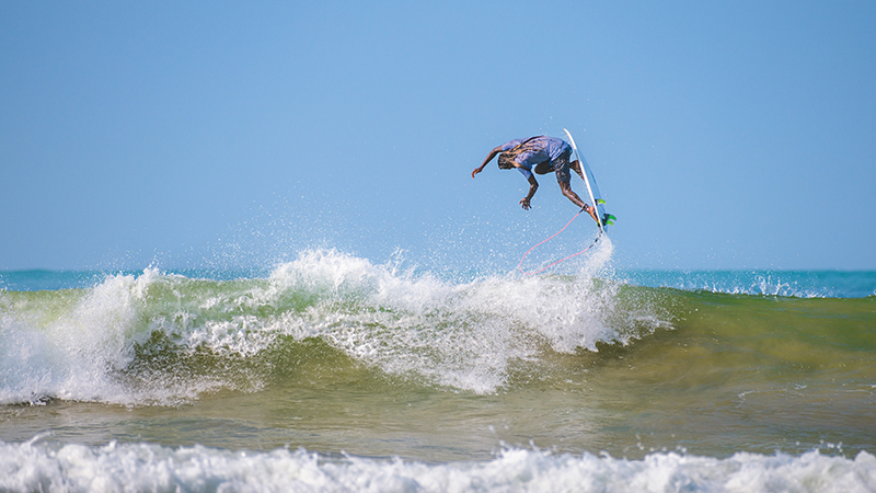 Surfer in mid-air performing a trick above a breaking wave under a clear blue sky.